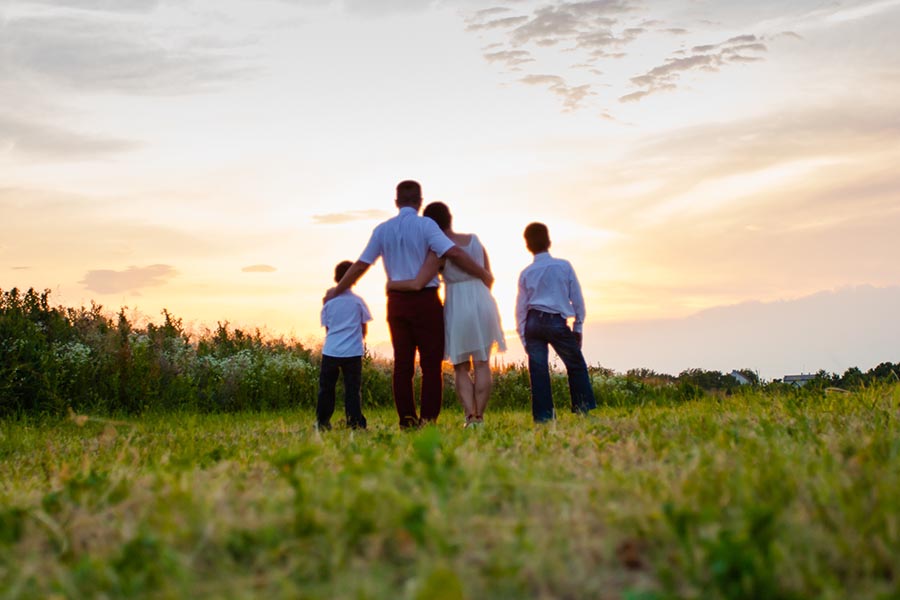 About Our Agency - Family Stands in a Green Field With Their Arms Around One Another, Watching a Golden Sunset