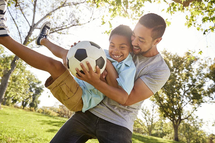 Personal Insurance - Father Scoops up His Son as They Laugh and Play Soccer in Their Large Back Yard With Trees and Blue Sky Overhead